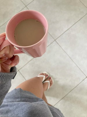 Person in gray sweater and white sandals holding pink mug, standing on tiled floor.