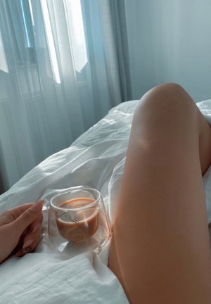 Person's legs on bed holding coffee cup near window.
