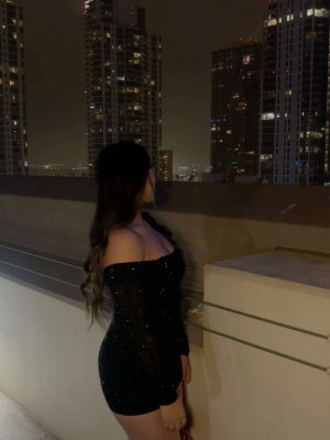 Woman in black sequined off-shoulder dress on rooftop at night, city skyline behind.