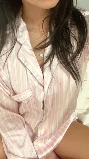 Woman in pink striped pajama shirt, sitting indoors, dark hair, necklace visible.