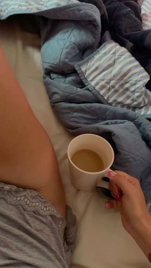 Woman in gray lace underwear holding coffee mug in bed, covered by blue quilt.