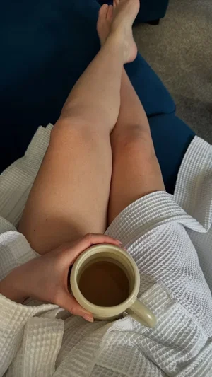 Woman in white robe holding mug, legs crossed on blue couch.
