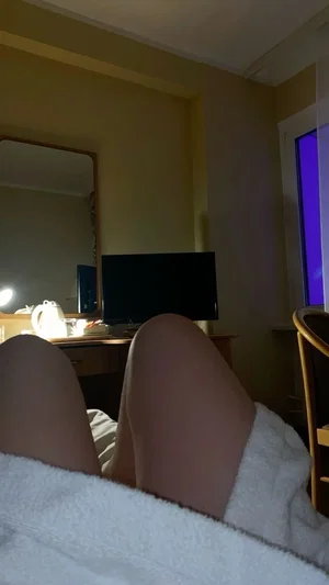 Person lying in bed with legs visible, covered by white blanket, in a bedroom with mirror and TV.
