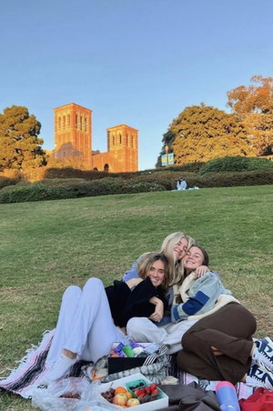 Three women picnic on grass near campus in casual clothes.