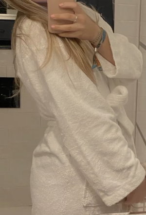 Woman with long blonde hair in white bathrobe taking bathroom selfie.