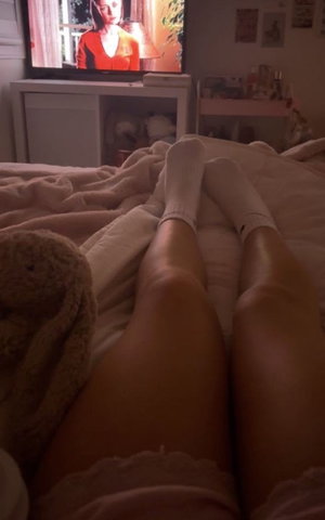 Legs in bed with white socks and pink lace, TV on in background.