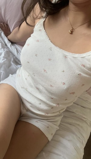 Woman in white floral tank top and shorts sitting on bed, wearing gold necklace.