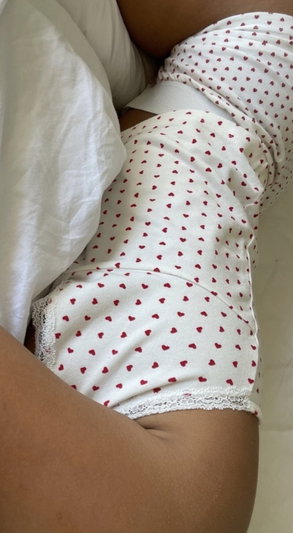 Person in white underwear with red hearts, lying on bed with white sheets.