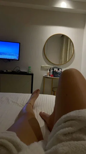 Person in white robe sitting on bed, legs visible, room with TV and mirror.