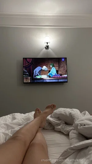 Person's bare legs on bed, watching anime on TV in bedroom.