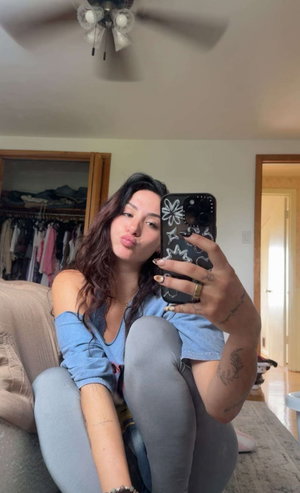 Woman with long dark hair in blue off-shoulder top and gray leggings, sitting on couch, taking