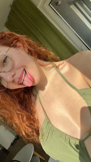 Woman with red curly hair and glasses wearing green top, sticking tongue out indoors.