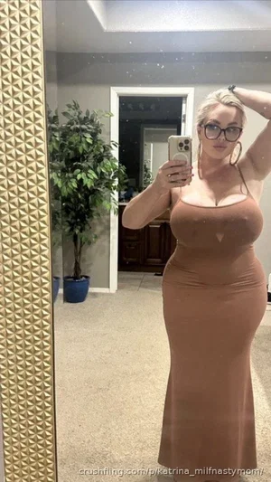 Woman in a brown form-fitting dress takes mirror selfie in a room with plants and a vanity.