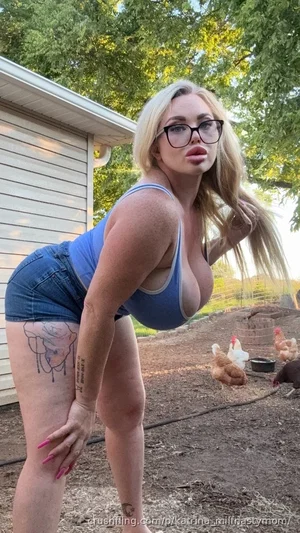 Woman with blonde hair and glasses in blue top and denim shorts posing outdoors near chickens.