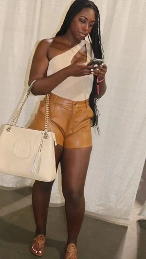 Woman with long braids in one-shoulder top and brown shorts, holding phone and purse indoors.