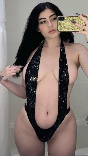 Woman with long dark hair in black sequined bodysuit taking mirror selfie in bathroom.