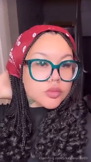 Woman with braided hair, red bandana, green glasses, black top, indoors.