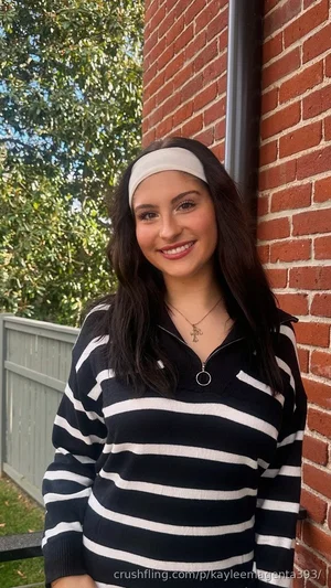 Woman with long dark hair wearing striped sweater and headband, standing by brick wall outdoors.