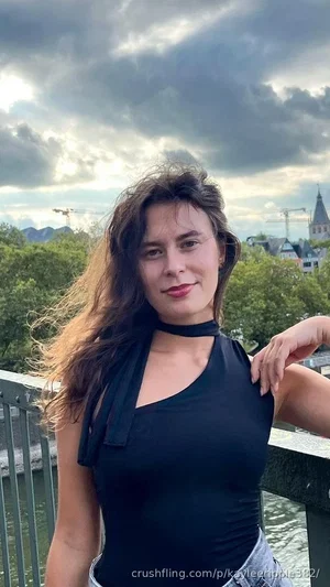 Woman with long dark hair in black top and jeans, standing on bridge with city and clouds behind.