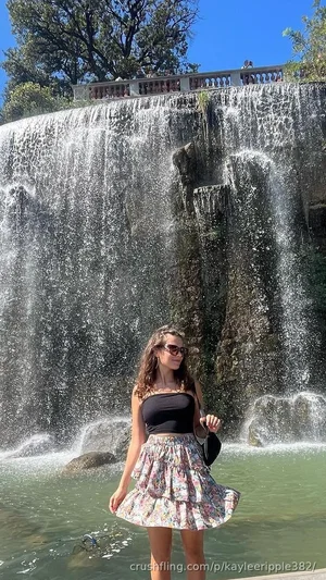 Woman in black crop top and floral skirt standing by waterfall in outdoor setting.