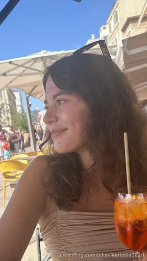 Woman with dark curly hair in a beige strapless top, holding a drink outdoors.