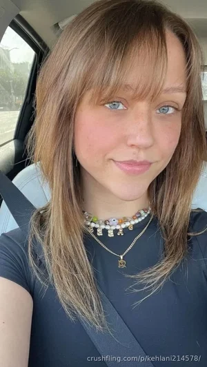 Woman with light brown hair and blue eyes wearing a dark top and necklaces, sitting in a car.
