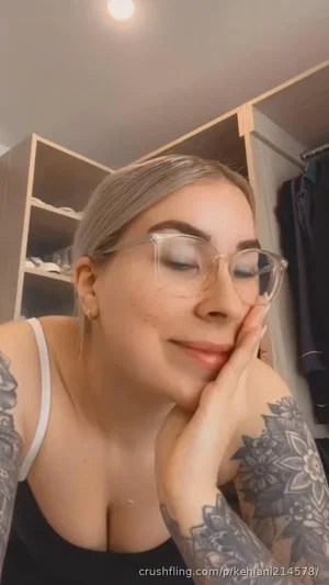 Woman with tattoos and glasses in a black top, indoors near closet.