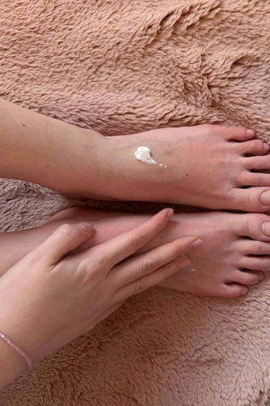 Hands applying cream to feet on pink fluffy surface.