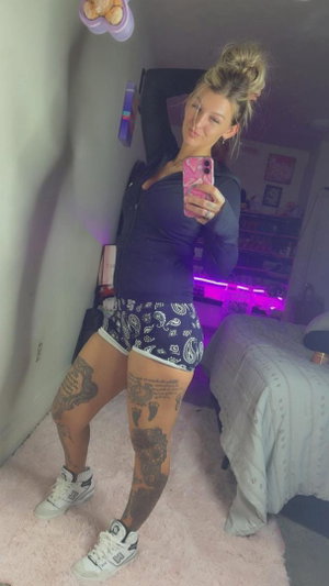 Blonde woman with tattoos taking mirror selfie in bedroom.