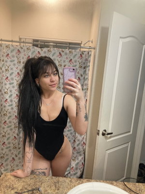 Woman in black one-piece swimsuit takes mirror selfie in bathroom with patterned shower curtain.