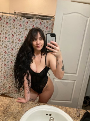 Woman with long dark hair and tattoos wears black lingerie in bathroom mirror selfie.