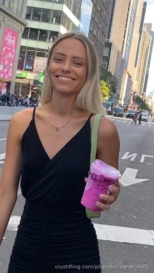 Blonde woman in black dress holding pink drink on city street.