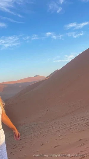 Woman in light clothing standing on a sand dune under a clear blue sky.