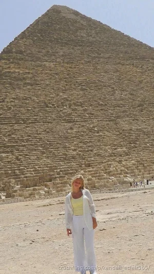 Woman in white pants and light top standing in front of a large pyramid in a desert setting.