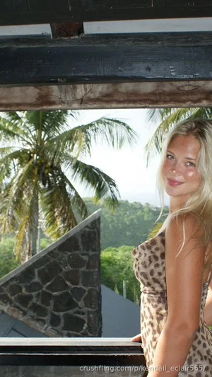 Blonde woman in leopard print dress standing on balcony with palm trees and green hills in