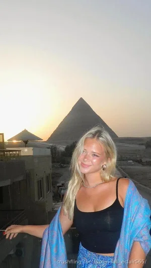 Blonde woman in black top and blue wrap, standing outdoors near pyramids at sunset.