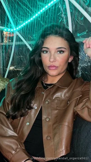 Woman with long dark hair in brown leather jacket and black top, posing indoors with green lighting.