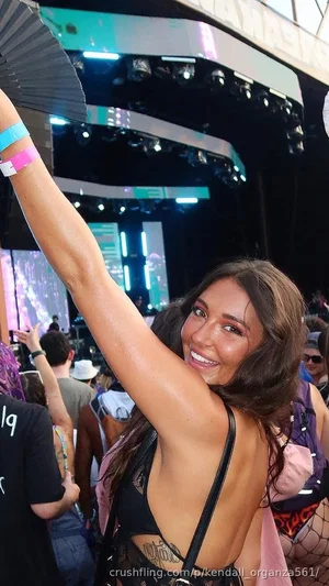 Woman with long dark hair in black lace top at outdoor event, smiling with arm raised.