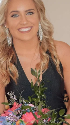 Blonde woman in black halter dress holding colorful flowers, smiling at camera.