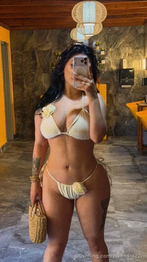 Woman in beige bikini with floral details, holding woven bag, taking mirror selfie in tiled room.
