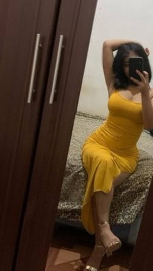Woman in yellow dress sits on bed, taking mirror selfie in bedroom.