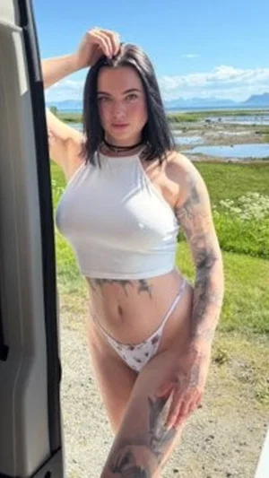 Woman with tattoos in white crop top and bikini bottoms standing outdoors near water.