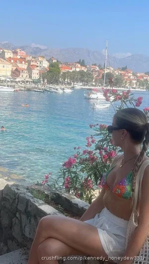 Woman in floral bikini top and white shorts sits by seaside, facing water with town and boats in