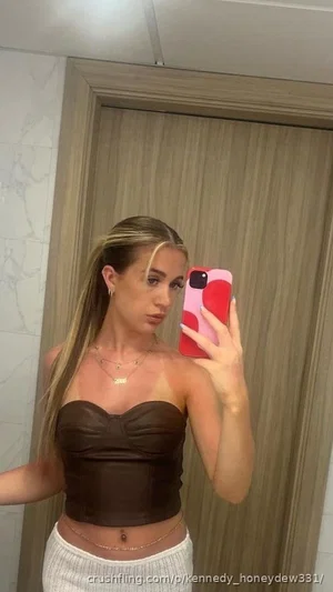 Woman with blonde hair in brown top and white skirt takes mirror selfie in bathroom.