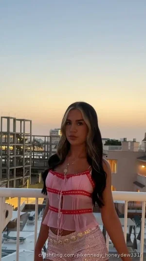 Woman in pink sheer top and sequined shorts on balcony at sunset.