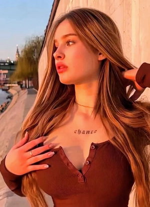 Woman with long blonde hair in brown top, standing outdoors near water, hand on chest.