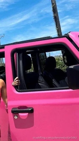 Woman in black top standing by bright pink jeep, hand on door, outdoor setting.