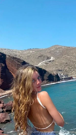 Woman with curly blonde hair in white bikini top by the ocean, rocky cliffs in background.