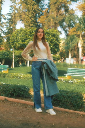 Woman with glasses in white top and jeans holding jacket in park.
