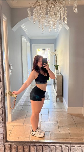 Woman in black sports bra and shorts takes mirror selfie in hallway with chandelier.